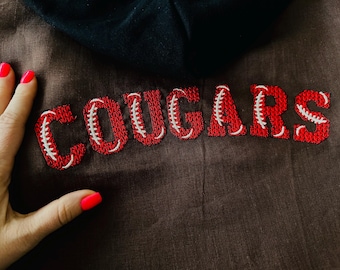 May include: A brown fabric garment with the word "COUGARS" embroidered in red with white stitching, resembling a baseball.
