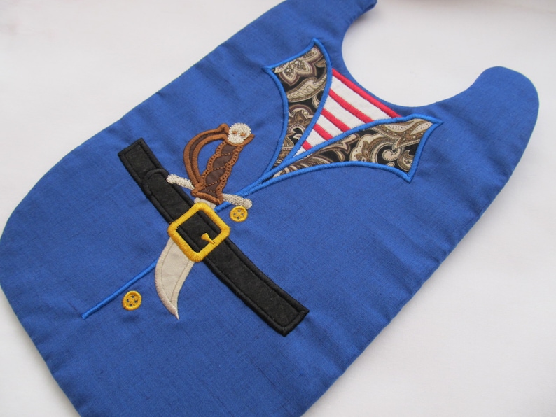 May include: A blue fabric bib with an embroidered design of a pirate costume, including a sword, belt, and shirt collar.