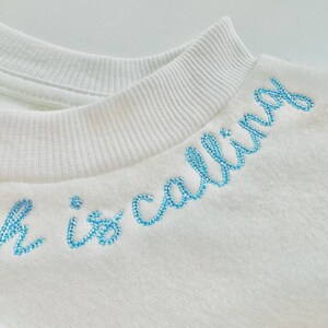May include: Close-up of a white sweatshirt with a ribbed collar. The phrase "witch is calling" is embroidered in light blue cursive across the chest. The fabric appears soft and textured.