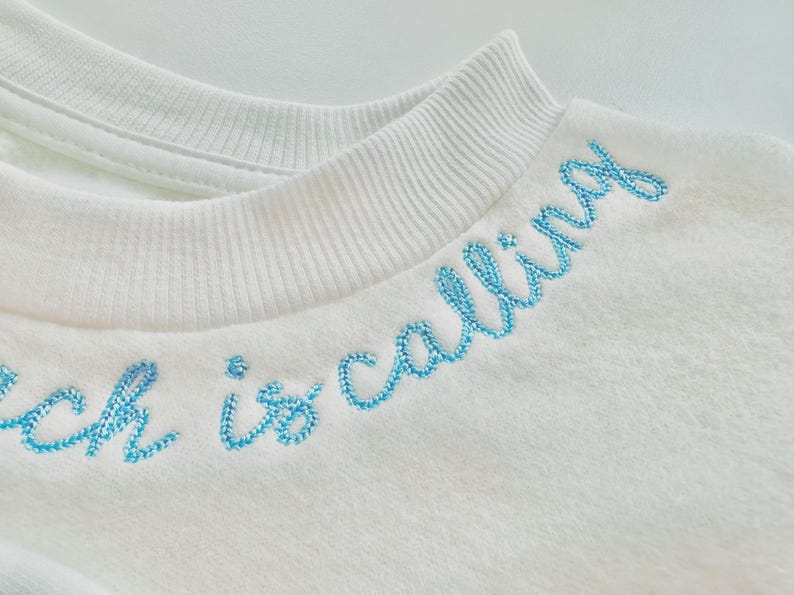 May include: Close-up of a white sweatshirt with a ribbed collar. The phrase "witch is calling" is embroidered in light blue cursive across the chest. The fabric appears soft and textured.