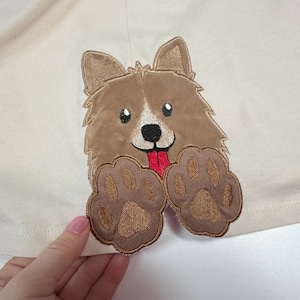 Op de afbeelding: Een bruine geborduurde hond met een rode tong die uitsteekt, met twee bruine poot afdrukken op een beige stoffen achtergrond.