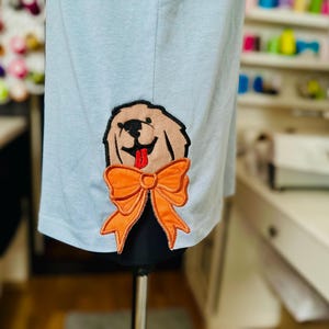 以下が含まれることがあります： オレンジ色の蝶ネクタイをした犬の刺繍パッチが付いた薄いブルーのTシャツ