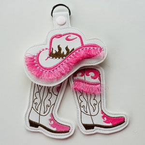 May include: A pink and white cowboy hat and boots keychain. The hat has a brown embroidered design and pink fringe. The boots have white embroidery and pink fringe.