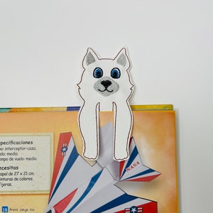 May include: White felt bookmark with an embroidered white dog with blue eyes. The bookmark is placed on a book with a paper airplane on the cover. The airplane is red, white, and blue with the word "NASA" printed on it.