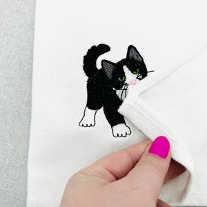 May include: White cloth with a black and white embroidered cat design. The cat has green eyes and a pink nose. The cloth has a herringbone pattern.