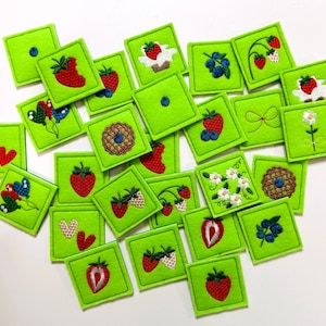 Könnte beinhalten: Ein Set mit 20 grünen Filzquadraten mit verschiedenen gestickten Designs, darunter Erdbeeren, Blaubeeren, Blumen und eine Torte. Die Quadrate eignen sich perfekt für ein Bastelprojekt oder ein Spiel.