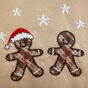 May include: Two gingerbread people embroidered on a beige fabric. One gingerbread person is wearing a red and white Santa hat. Both gingerbread people have brown and white plaid patterns on their bodies. There are white snowflakes embroidered around the gingerbread people.