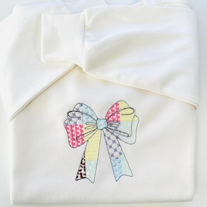 May include: A white sweatshirt with a colorful embroidered bow. The bow is made up of different colored patches, including pink, yellow, blue, and brown.