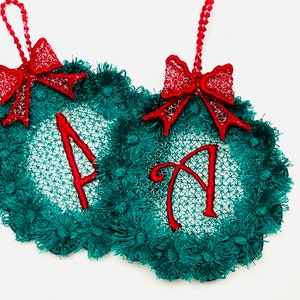 Peut inclure: Deux décorations de Noël brodées de couleur verte et rouge avec un motif de couronne et la lettre "A" au centre. Chaque décoration est surmontée d'un nœud rouge.