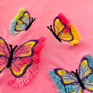 Puede incluir: Tela rosa con tres mariposas bordadas. Las mariposas son en tonos de rosa, amarillo, azul y verde con contornos negros. Las mariposas tienen bordes con flecos.
