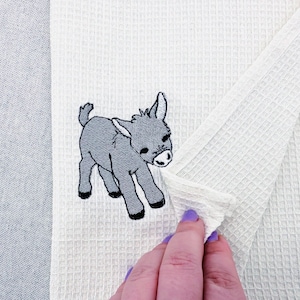 May include: White waffle-weave towel with an embroidered gray donkey design. The donkey is depicted in a walking pose with black hooves and a white muzzle. The towel is folded, revealing its textured surface.