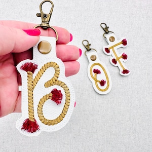 May include: White leather keyrings with gold rope-style embroidery forming the letters "B", "O", and "T". Each keyring has a red tassel and a bronze-coloured clasp. The keyrings are held by a person with pink nail polish.