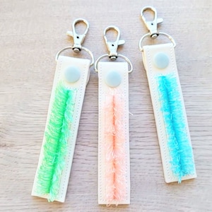 May include: Three white keyrings with different coloured fuzzy trim. The keyring on the left has green trim, the middle keyring has pink trim, and the keyring on the right has blue trim.