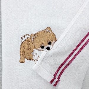 May include: White towel featuring an embroidered brown Pomeranian dog. The towel has a red striped border. The dog is lying down, with its head resting on the towel. The towel is made of a soft, textured fabric.