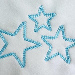 Puede incluir: Tres estrellas bordadas de diferentes tamaños se muestran sobre un fondo blanco. Las estrellas están delineadas con una puntada azul claro, creando un efecto texturizado. El diseño es simple y limpio.