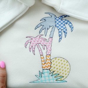 May include: White sweatshirt with a colorful embroidered palm tree design. The embroidery features a pink, blue, and yellow palm tree with a sun and blue water. The design is on the front of the sweatshirt.