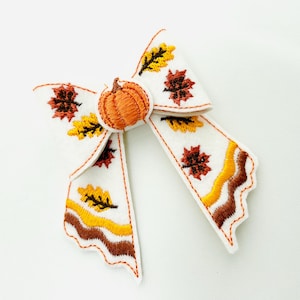May include: A white fabric bow with embroidered autumn leaves and a pumpkin in the center. The bow features orange, yellow, and brown leaf designs, with a scalloped edge. The pumpkin is orange.
