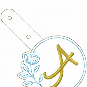 May include: A gold embroidered letter A with a blue flower design in a circle. The design is on a white fabric background.