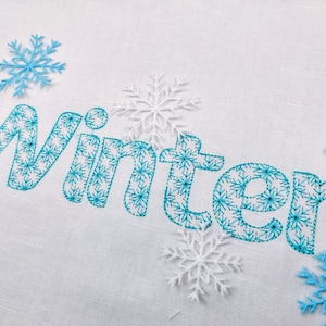 Könnte beinhalten: Weißer Stoff mit dem Wort "Winter" in blauen Schneeflockenmustern gestickt. Das Wort ist von blauen und weißen Schneeflocken umgeben.