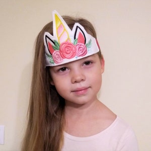 Puede incluir: Una diadema blanca con un cuerno de unicornio, orejas y flores rosas y rojas bordadas. La diadema la lleva puesta una niña con el pelo largo y rubio.