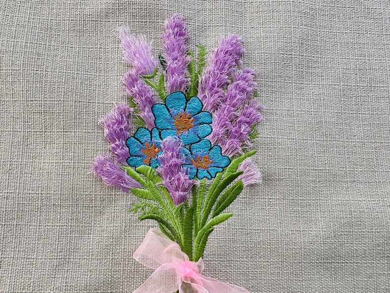 May include: Embroidered lavender flowers with blue centers, tied with a pink ribbon on a linen fabric background.