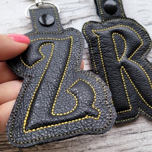 May include: Two black leather keychains with yellow stitching. The keychains are shaped like the letters "Z" and "R".