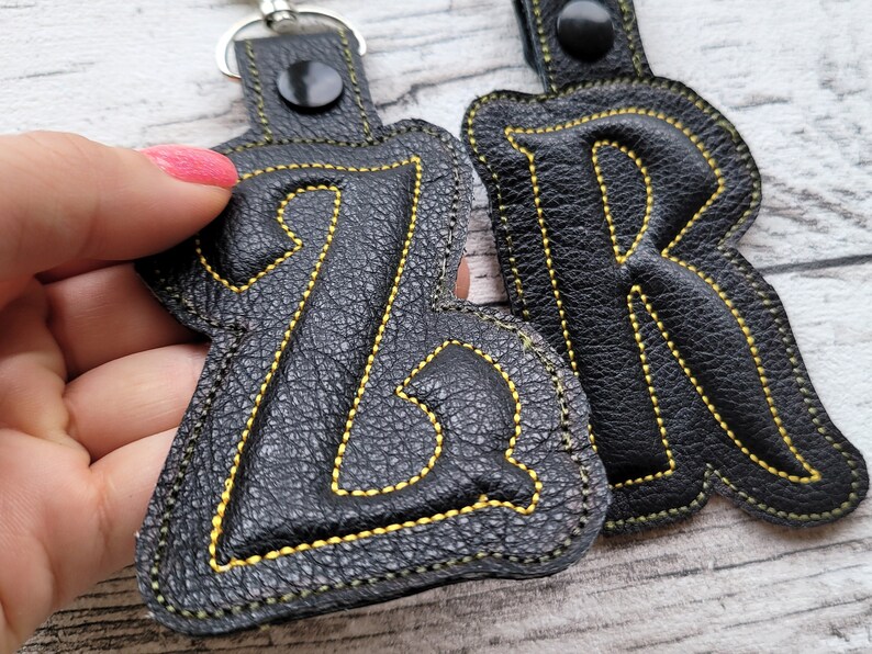 May include: Two black leather keychains with yellow stitching. The keychains are shaped like the letters "Z" and "R".