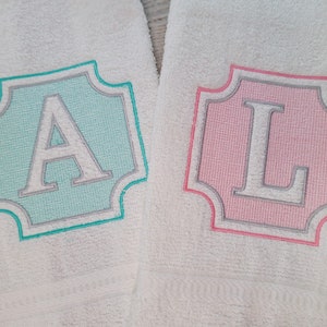 May include: Two white hand towels with embroidered initial designs. One towel features a light blue background with a gray border and the letter "A". The other towel has a pink background with a gray border and the letter "L".