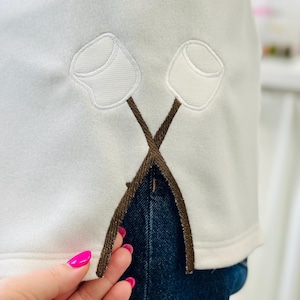 Op de afbeelding: Witte sweatshirt met een geborduurd ontwerp van twee marshmallows op gekruiste stokken. De marshmallows zijn wit met een lichtbruine omtrek, en de stokken zijn bruin. De sweatshirt wordt gecombineerd met een blauwe jeans.