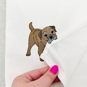 May include: A white tea towel featuring an embroidered brown Border Terrier dog with a grey muzzle. The fabric has a subtle herringbone weave pattern. The embroidery shows the dog standing in profile with its tail pointed upward.