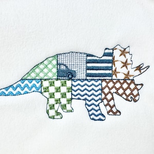 May include: Embroidered triceratops design in various patchwork patterns. The dinosaur features blue, green, and brown patches with a small car detail. The design is on a white background.