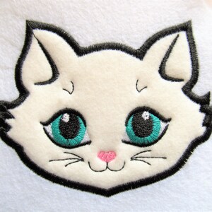 Puede incluir: Cara de gato blanco bordada con contorno negro, ojos azul turquesa y nariz rosa.