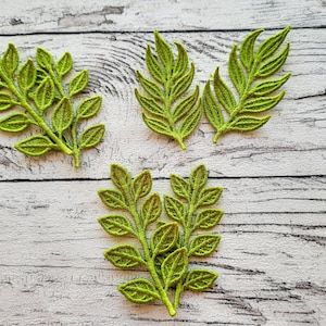 Puede incluir: Primer plano de varios ramitos de hojas bordadas de color verde brillante sobre una superficie de madera blanca desgastada. Las hojas tienen costuras detalladas, creando una apariencia texturizada. Los ramitos varían en tamaño y forma, sugiriendo un tema botánico natural.