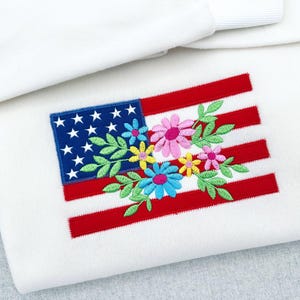 以下が含まれることがあります： 白い生地のアイテムで、アメリカ国旗のデザインが刺繍されています。国旗の青い部分には白い星があり、赤いストライプは、ピンク、青、黄色の花と緑の葉のフラワーアレンジメントで飾られています。アイテムは折りたたまれています。