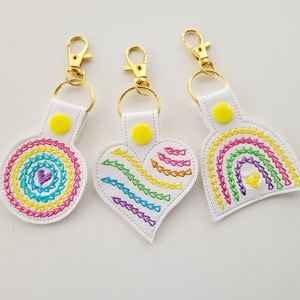 Puede incluir: Tres llaveros blancos con herrajes dorados. Cada llavero presenta un diseño bordado diferente: un círculo arcoíris, un corazón arcoíris y un arcoíris. Los diseños están cosidos con vibrantes colores del arcoíris.