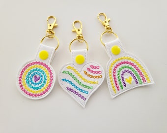 May include: Three white keyrings with gold-coloured clasps. Each keyring has a different embroidered design: a rainbow circle, a rainbow heart, and a rainbow arch. The designs are stitched with vibrant rainbow colours.