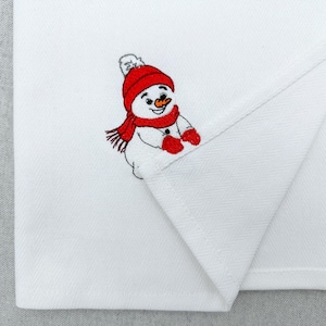 May include: A white kitchen towel featuring an embroidered snowman wearing a bright red knit hat, a matching red scarf, and red mittens. The snowman has a small orange carrot nose and a smiling expression, positioned on the folded corner.
