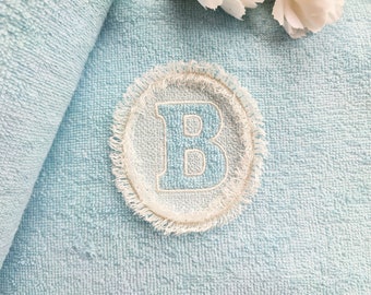 May include: A light blue towel with a white embroidered circle containing the letter "B" in a light blue color.