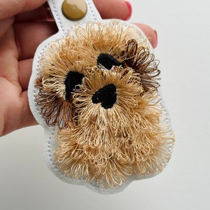 May include: A white leather keychain featuring an embroidered dog face in shades of brown and gold. The dog has black eyes and a brown nose.