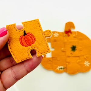 May include: A felt pumpkin puzzle with an orange and black pumpkin design. One puzzle piece is held in a hand, showing an embroidered orange pumpkin. The completed puzzle is in the background, with a bow and floral accents.