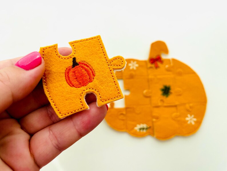 May include: A felt pumpkin puzzle with an orange and black pumpkin design. One puzzle piece is held in a hand, showing an embroidered orange pumpkin. The completed puzzle is in the background, with a bow and floral accents.