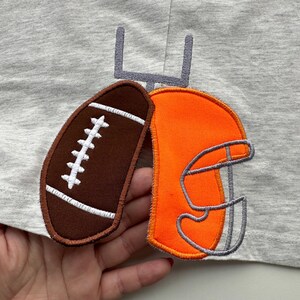 May include: A gray t-shirt featuring embroidered football-themed patches. The patches include a brown football with white stitching, an orange football helmet, and a gray goalpost. The design is likely for a child's apparel.