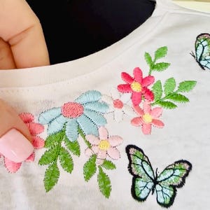 Puede incluir: Camiseta blanca con flores bordadas y una mariposa. Las flores son rosas, azules y amarillas. La mariposa es verde y azul.