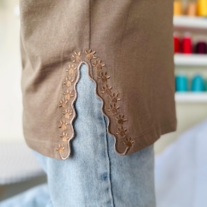 Puede incluir: Una camisa marrón con un diseño bordado decorativo en la costura lateral. El bordado es un borde festoneado con pequeños motivos florales.