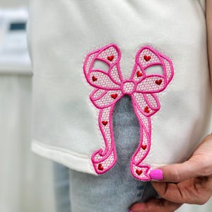 Puede incluir: Prenda blanca con un gran bordado de lazo rosa. El lazo presenta un delicado diseño de encaje con pequeños acentos de corazón rojo. Las cintas del lazo se extienden hacia abajo, agregando un toque decorativo a la ropa.