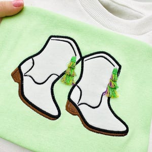 Puede incluir: Una camiseta blanca con fondo verde que presenta un diseño bordado de dos botas de vaquero blancas con borlas verdes y moradas.