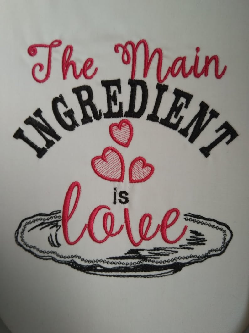 K&ouml;nnte beinhalten: Besticktes K&uuml;chentuch mit dem Text "The main ingredient is love" in schwarzer und rosa Schrift. Drei rosa Herzen befinden sich &uuml;ber dem Text, und eine schwarze Umrandung einer Servierplatte befindet sich unter dem Text.