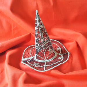 Puede incluir: Un sombrero de bruja negro y plateado con un cono puntiagudo y un diseño de estrella. El sombrero está hecho de encaje y está sobre un fondo de tela roja.