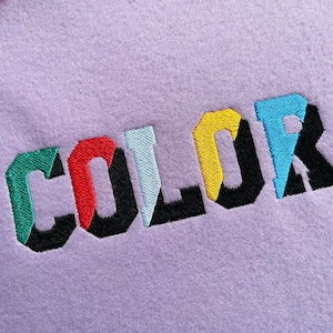 May include: Embroidered "COLOR" in a multi-coloured font on a purple fabric background.