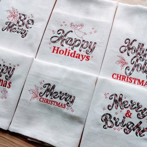 May include: Set of six white kitchen towels with red and black checked embroidered Christmas sayings. The sayings include "Be Merry", "Happy Holidays", "Holly Jolly Christmas", "Happy Christmas", "Merry Christmas", and "Merry & Bright".